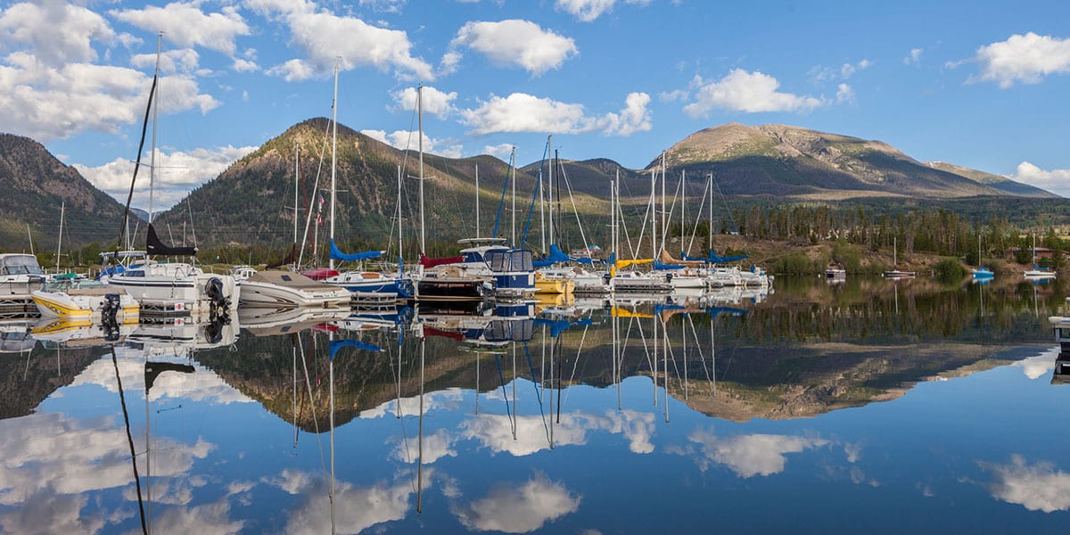 On a clear day, boats docked in the marina and mountains in the background are reflected perfectly by calm water.