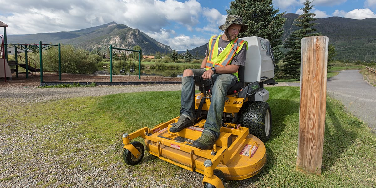 A public works employee cuts grass a Frisco playground using a riding mower
