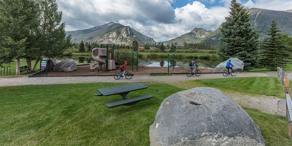 Three bikers riding on gravel path through Meadow Creek Park with playground and pond in background